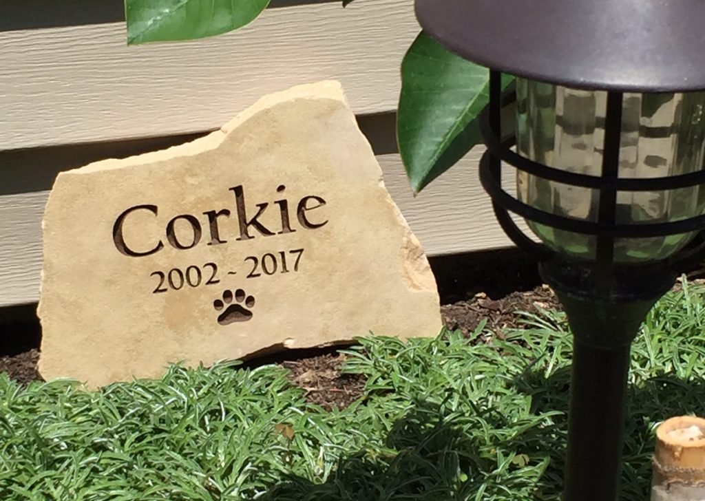 A memorial stone engraved with the name 'Corkie', the years 2002 to 2017, and a paw print, surrounded by green plants and next to a garden lantern.