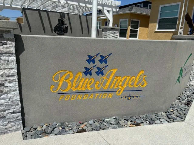 Sign for Blue & Angels Foundation with yellow text and three blue angel silhouettes above, located outdoors on a concrete wall surrounded by gravel, with a building and sky in the background.