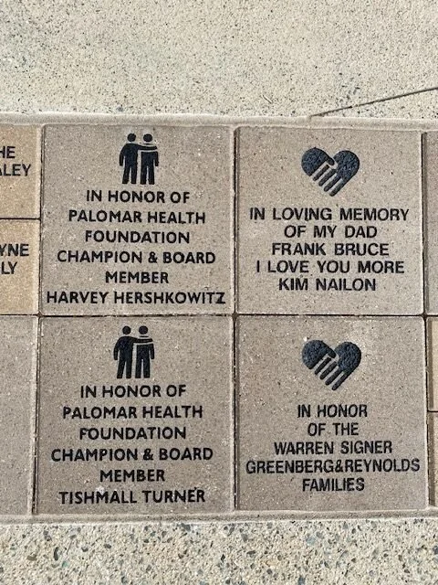 Four engraved bricks on the ground with memorial inscriptions and symbols. The top left brick honors Harvey Hershkowitz of the Palomar Health Foundation; the top right brick is a loving memory of Frank Bruce by Kim Nailon. The bottom left brick honor