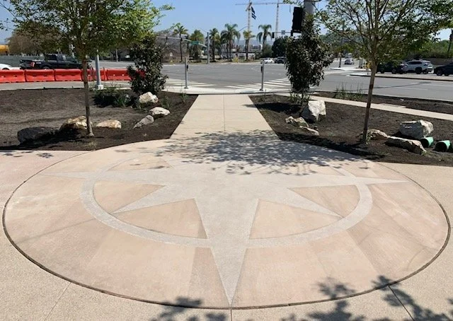 Sidewalk with a star and compass rose design at the center, flanked by small trees and rocks, with a street and cars in the background.