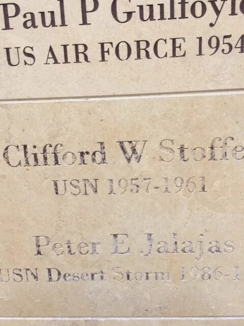 Close-up of a beige memorial plaque with black engraved text honoring military personnel, including Paul P. Gulloyo, Clifford W. Stoffel, and Peter E. Jajak, with their service details.