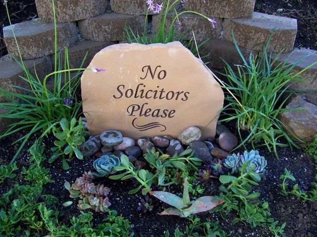 A decorative garden stone with the inscription 'No Solicitors Please' surrounded by green plants and succulents in a garden bed.