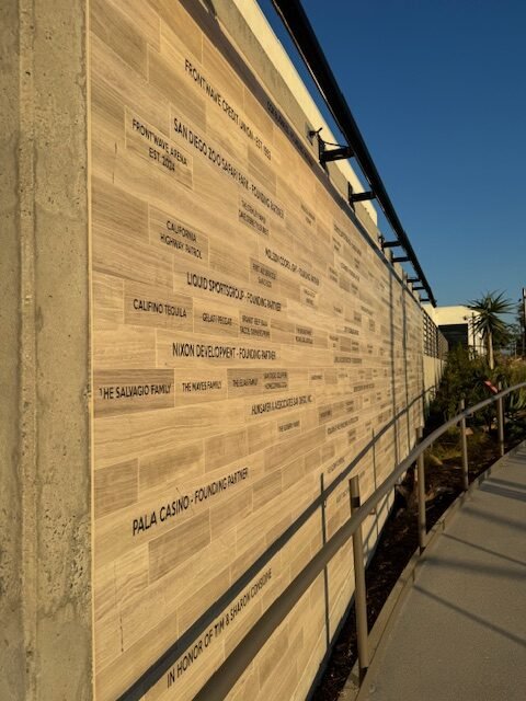 Photo of a wooden wall with engraved names of donors or supporters, next to a sidewalk and some plants, under a clear blue sky.