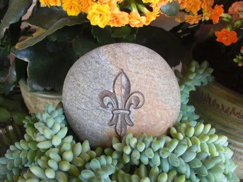 A round gray stone with a carved fleur-de-lis symbol, surrounded by green succulent plants, and part of a flower arrangement with orange and yellow flowers in the background.