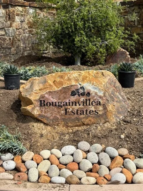A large decorative rock with the inscription 'Bougainvillea Estates' and a bougainvillea flower graphic, surrounded by small rocks, potted succulents, and green bushes in a landscaped garden.