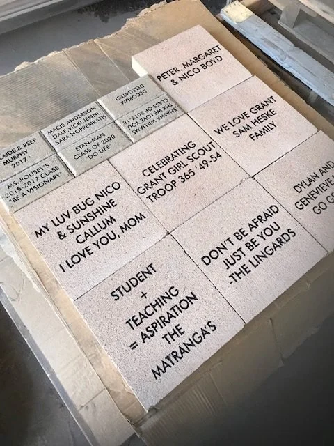A cardboard box contains several ceramic tiles with handwritten messages and names, including references to loved ones, family, and inspirational quotes.