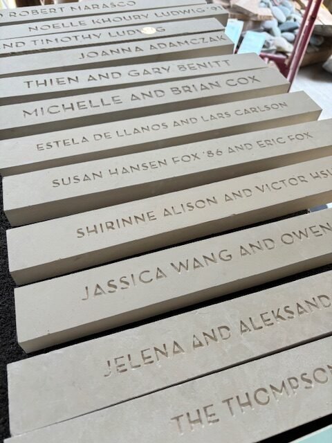 A row of beige plaques with engraved names, including Robert Narasco, Noelle Khoury Ludwig, Joanna Adamczyk, & Gary Benitt, with additional names partially visible, displayed on a black surface.