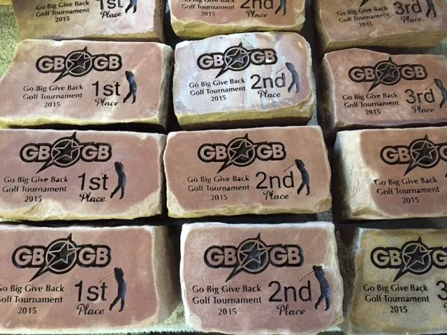 Multiple rocks with engraved logos and text for the 2015 Go Big Give Back Golf Tournament, showing placements 1st, 2nd, and 3rd.