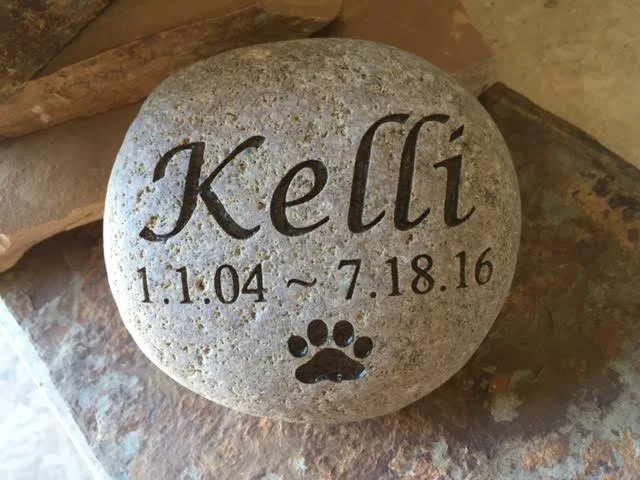 A decorative stone engraved with the name 'Kelli', a birth date and death date, and a paw print symbol.