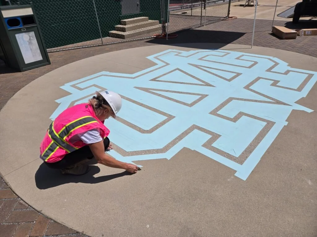 A worker in a white helmet and pink safety vest is drawing a large hopscotch pattern on a circular cement sidewalk using chalk.