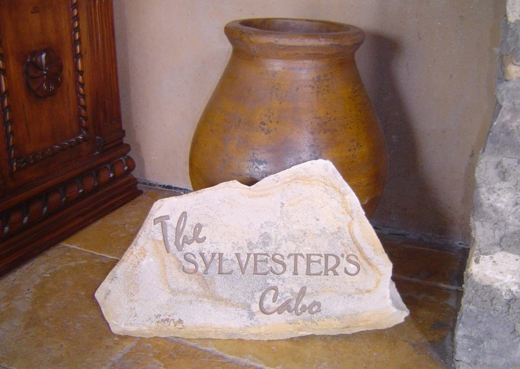 A sign made of stone with the words "The Sylvester's Cabo" engraved on it, placed on a tiled floor next to a large clay vase and wooden furniture.