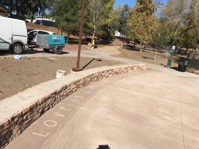 A curved concrete driveway with the word 'LOVE' and the number '104' painted in white along its edge, a stone retaining wall, a small tree, a blue portable generator, a white van, and a trash can in a park area with trees in the background.
