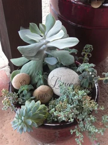 Decorative arrangement of succulents and stones with engraved words 'joy,' 'faith,' and 'peace,' in a red pot.