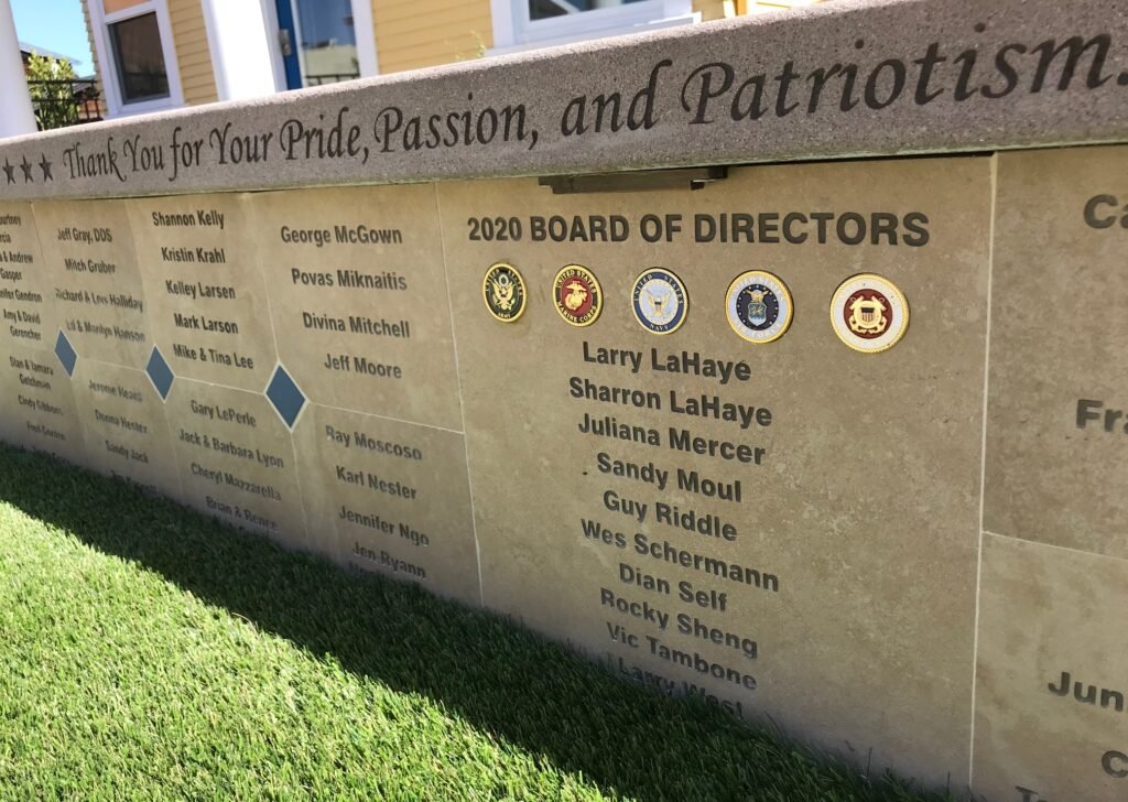 A commemorative stone wall displaying the 2020 Board of Directors and various logos, with a message thanking for pride, passion, and patriotism, situated outdoors on grass.
