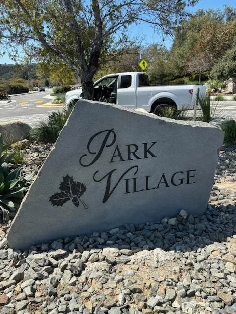 Large stone sign with the text "PARK VILLAGE" and a holly leaf logo, situated in a landscaped area with rocks. A white pickup truck is parked in the background near trees and a crosswalk.