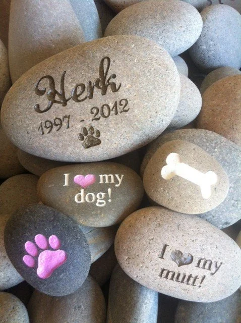 Collection of smooth stones with engraved or painted messages about dogs and pets, including the name 'Herk,' dates 1997-2012, and phrases like "I love my dog!" and "I love my mutt!" with paw print and bone illustrations.
