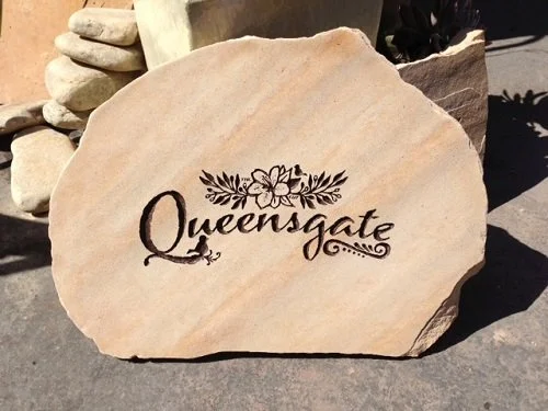 A large flat rock with the word 'Queensgate' and floral design engraved on it.