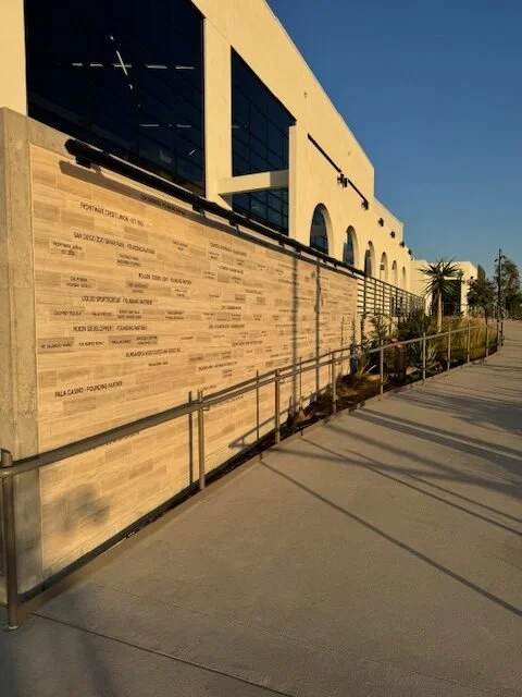 Exterior of a modern building with large windows and a stone wall with engraved text, a paved walkway, and palm trees in a sunny environment.