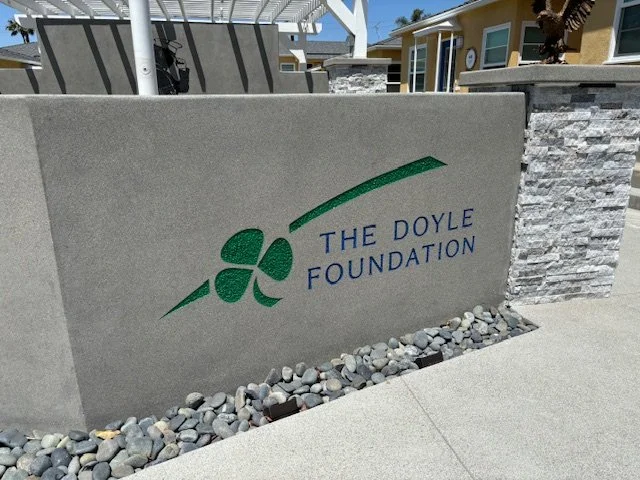 Concrete sign with a green clover logo and blue text reading 'The Doyle Foundation' in front of a residential area.
