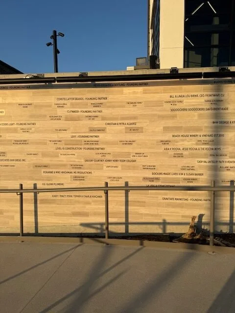 Large wooden wall with multiple lines of small black text, possibly names or business titles, next to a walkway with a metal railing, with a building and blue sky in the background.