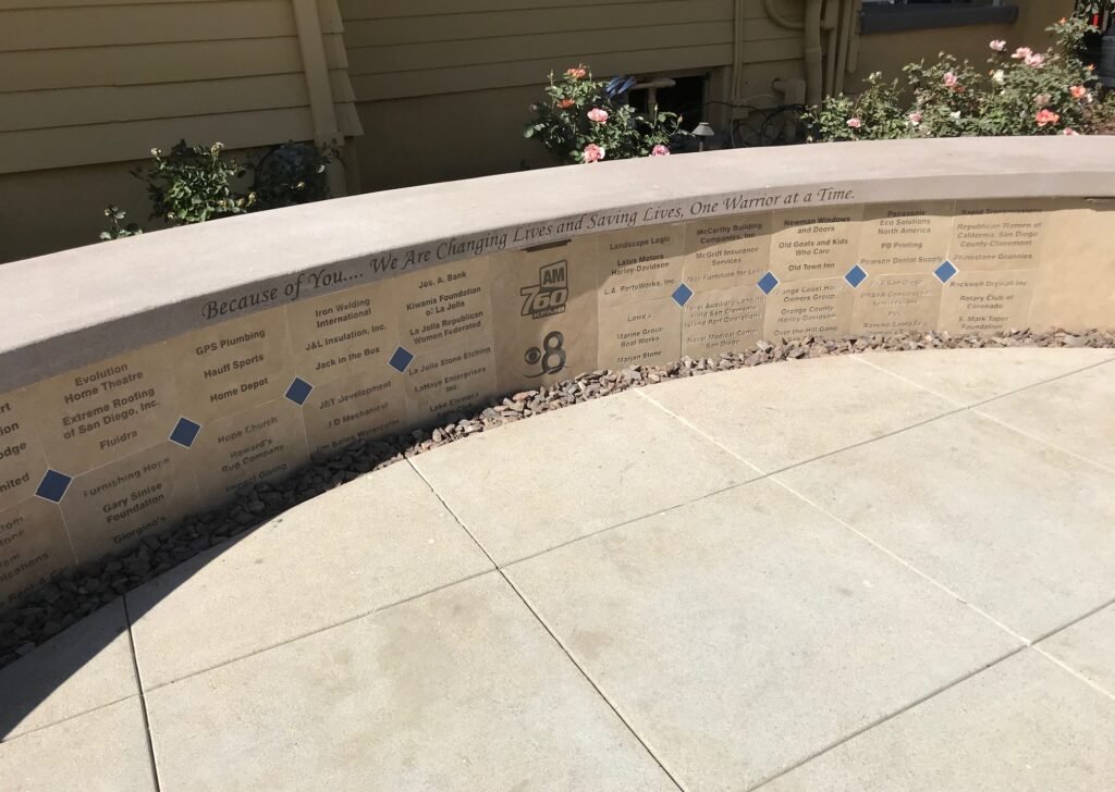 A curved stone memorial with engraved text and small blue tiles, surrounded by flowers and a sidewalk.