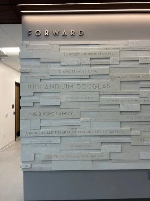 A wall with a list of names, including JUDI AND JIM DOUGLAS and THE BJORG FAMILY, under a sign that says "FORWARD" in a modern interior space.