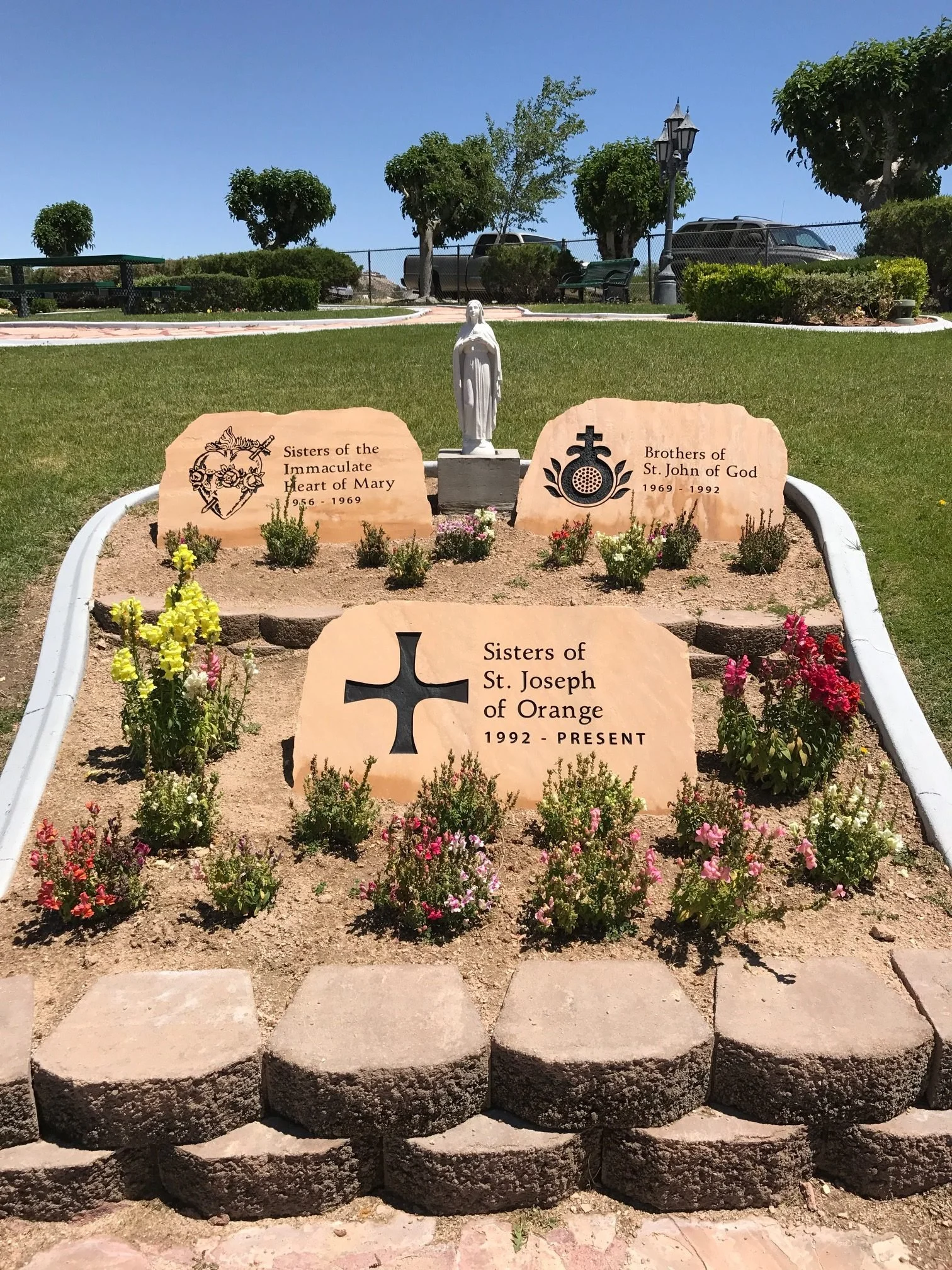 Gravel garden with pink and yellow flowers, three large stone memorials with inscriptions for religious groups, a statue of a woman, and a paved pathway in front.