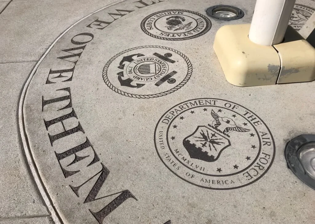 Close-up of a concrete surface with the emblems of the U.S. Marine Corps, U.S. Coast Guard, and the Department of the Air Force engraved on it, along with a portion of a metal pole and base.