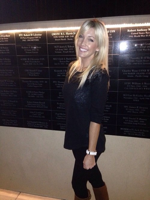 A woman with long blonde hair smiling, wearing a black top, black pants, and brown boots, standing indoors in front of a memorial wall with engraved names.