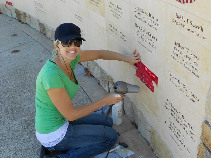 Smiling woman in green shirt and sunglasses installing a red memorial plaque on a stone wall at a memorial site, with her kneeling on the sidewalk.