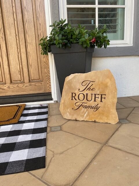 A decorative stone with the text "The ROUFF Family" engraved on it, placed on a stone doorstep next to a flower pot with green plants and a window with white shutters.