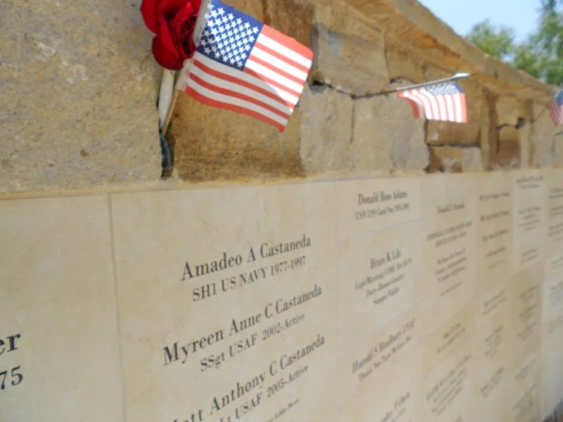 American flags hanging above engraved names of military personnel on a memorial wall in an outdoor setting.
