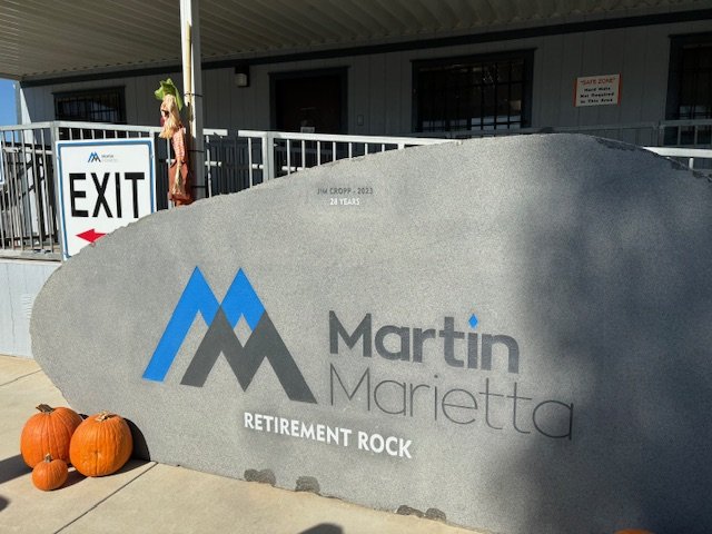 Large stone sign reading "Martin Marietta Retirement Rock" with a blue and gray logo, two pumpkins on the ground, and a woman standing near an exit sign in the background.