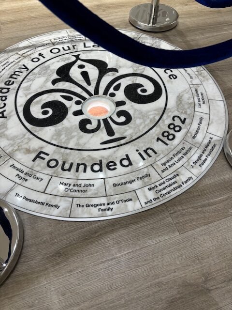 A circular table with a newspaper-like top displaying a logo, text indicating it was founded in 1882, and a list of family names, with part of a blue object hanging above it and a lamp in the background.