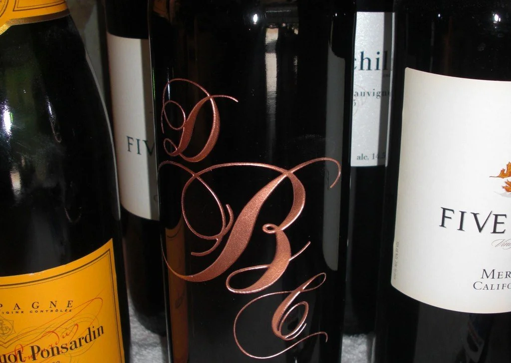 Close-up of wine bottles, one with a black label with copper-colored initials 'B' and 'L' in cursive, others with white labels and various colors.