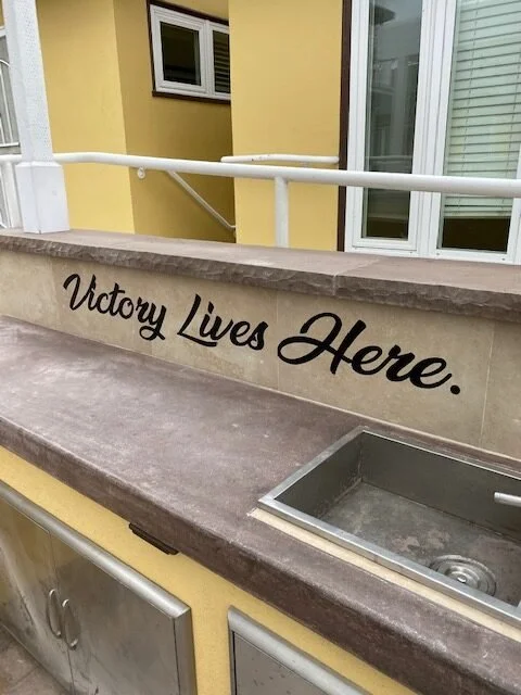 A yellow building exterior with a window, a white railing, and a stone counter with the phrase "Victory Lives Here." written on it in black cursive.