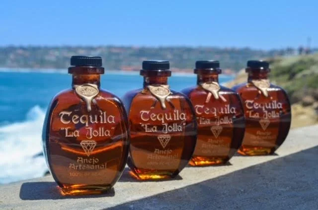 Four bottles of tequila labeled 'Tequila La Jolla' arranged in a row outdoors on a concrete surface, with a beach and ocean in the background on a sunny day.