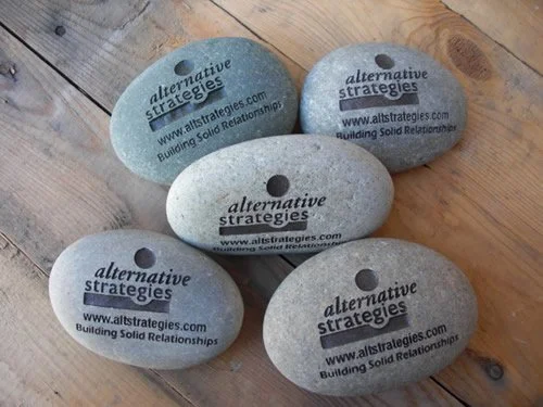Five smooth, rounded stones with printed text and website address, resting on a wooden surface.