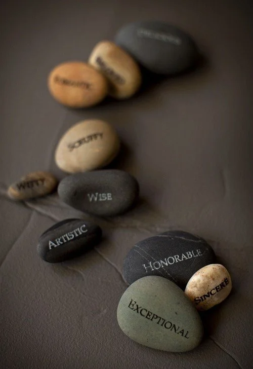 Colorful stones with words such as "WITTY," "WISDOM," "ARTISTIC," "HONORABLE," "SINCERE," and "EXCEPTIONAL" written on them, arranged on a dark surface.