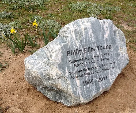 Gravestone with inscription for Philip Ellis Young, 1945-2011, amid flowers and grass.
