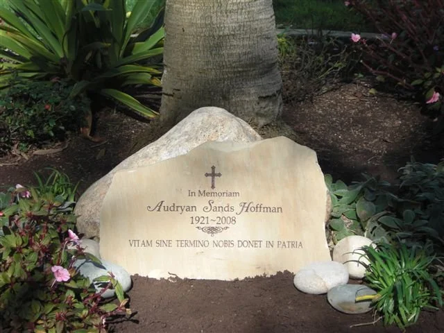 Memorial headstone for Audryan Sands Hoffman, 1921-2008, with a cross and a Latin phrase, surrounded by flowers and rocks in a garden.