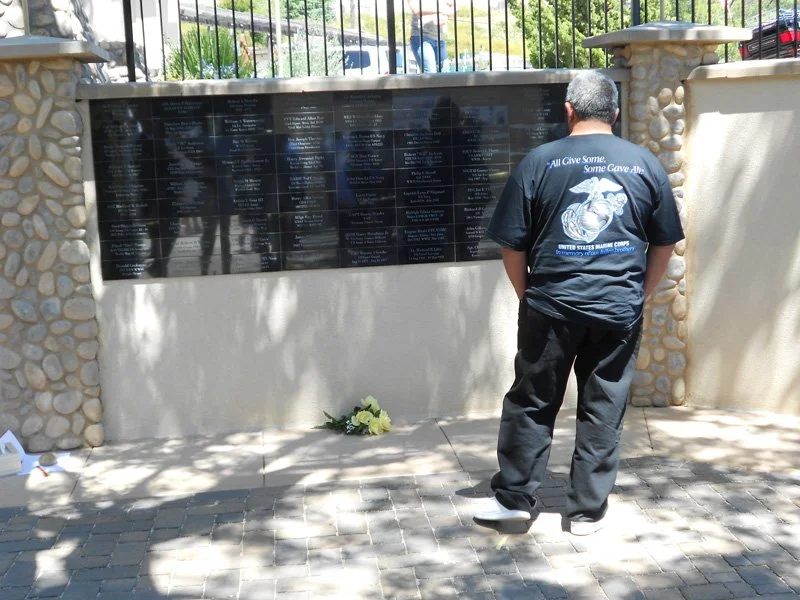 A man stands with his back to the camera, looking at a memorial wall with names inscribed on it. The man is wearing a dark t-shirt with a Marine Corps graphic and text, and dark pants. There is a small bouquet of flowers placed at the base of the wal