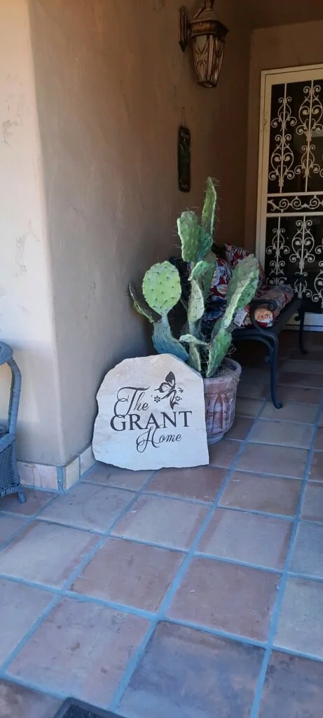 Potted cactus next to a decorative stone sign that reads "The Grant Home" with a butterfly graphic, on a tiled porch with a chair and a front door in the background.
