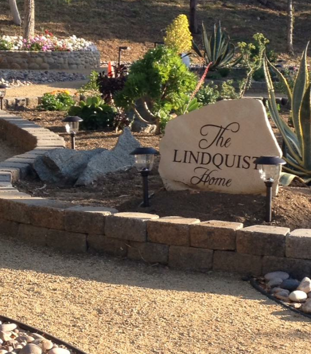 Garden with a large stone engraved with 'The Lindquist Home' surrounded by plants and small solar lights.