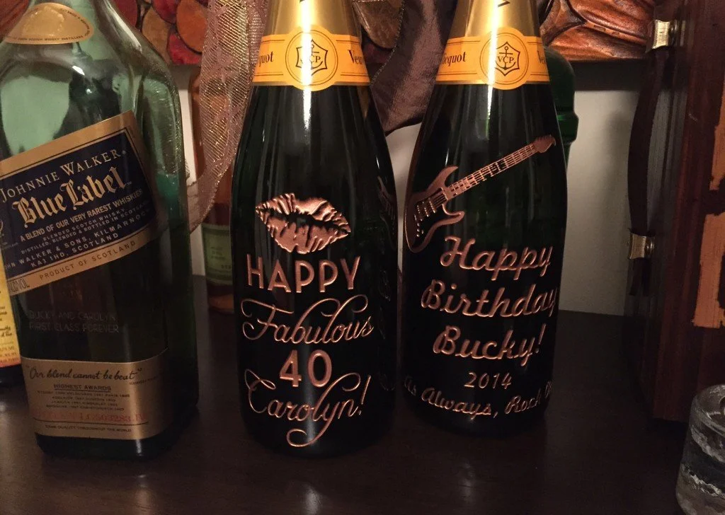 Two personalized black bottles with gold caps and custom labels are placed on a dark wooden surface. The left bottle's label reads 'HAPPY Fabulous 40 Carolyn!' with a lip graphic, and the right bottle's label reads 'Happy Birthday Bucky! 2014, Always