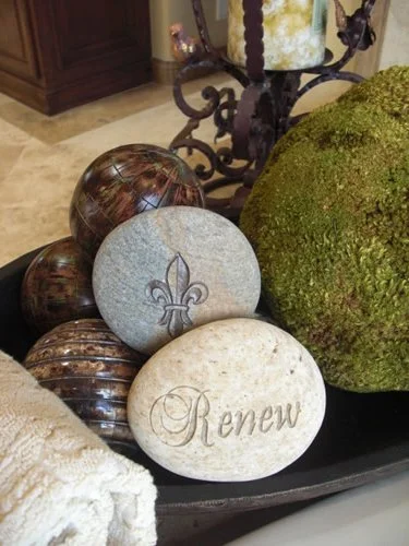 Decorative arrangement with painted and engraved stones, a moss-covered sphere, and a white rolled towel on a tray.