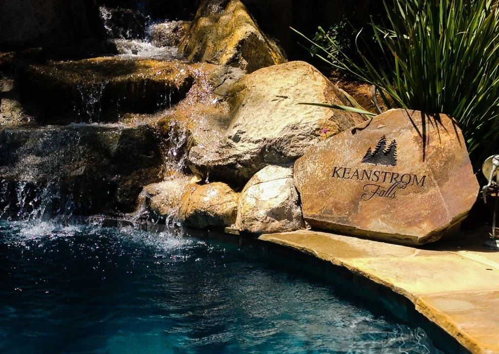 A pool with a small waterfall feature flowing over rocks, with a large engraved stone that reads 'Keenstrom Falls' and an illustration of trees.