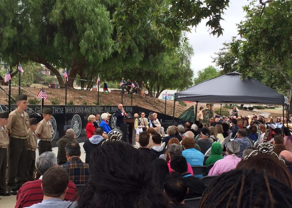 A large outdoor gathering with a stage, where people are seated under a black canopy and on chairs. Military personnel in uniform are standing on the left. An individual is speaking at a podium, and a black banner with white text and patriot flags is