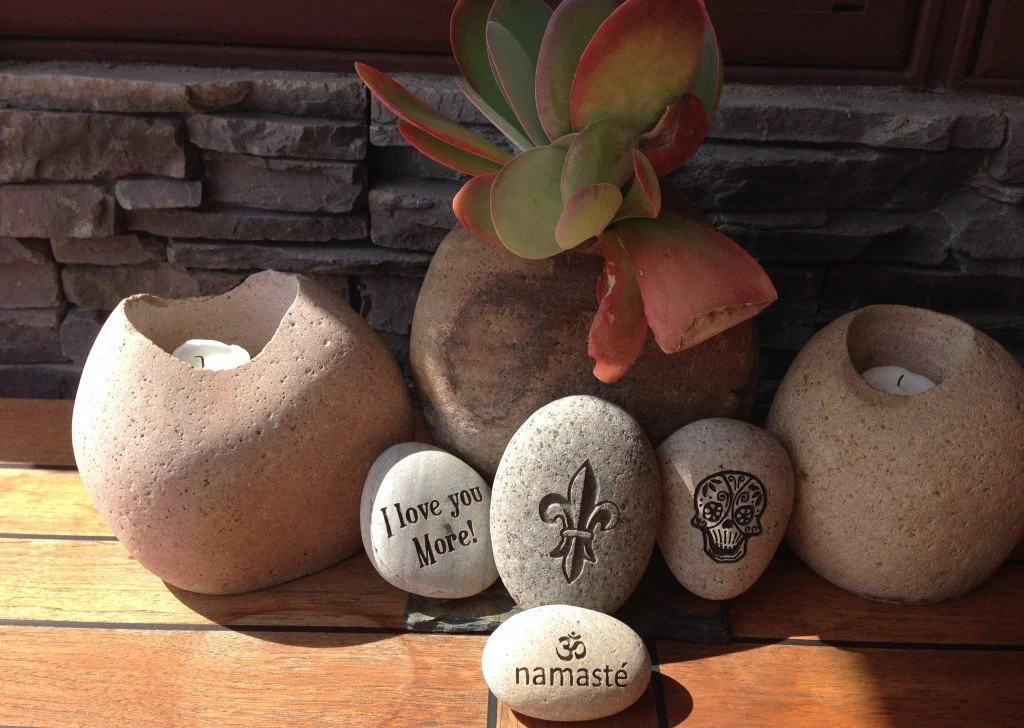 A collection of decorative stones, a succulent plant in a ceramic pot, and two candle holders with candles. One stone has the message "I love you more!" written on it, another has a fleur-de-lis symbol, one has a skull design, and the smallest stone 