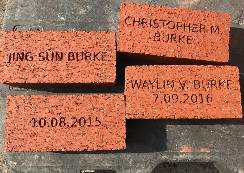 Four red bricks with engraved names and dates on a dark surface. The bricks have inscriptions: "Jing Sun Burke," "Christopher M. Burke," "Waylin V. Burke 7.09.2016," and "10.08.2015."